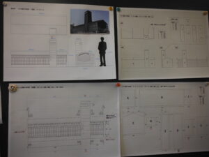 壁面に掲示された4枚の資料に、建物の外観写真やシルエットによる人物との比較図、そして各パーツの寸法や構造が細かく記された展開図や平面図が並んでいる写真