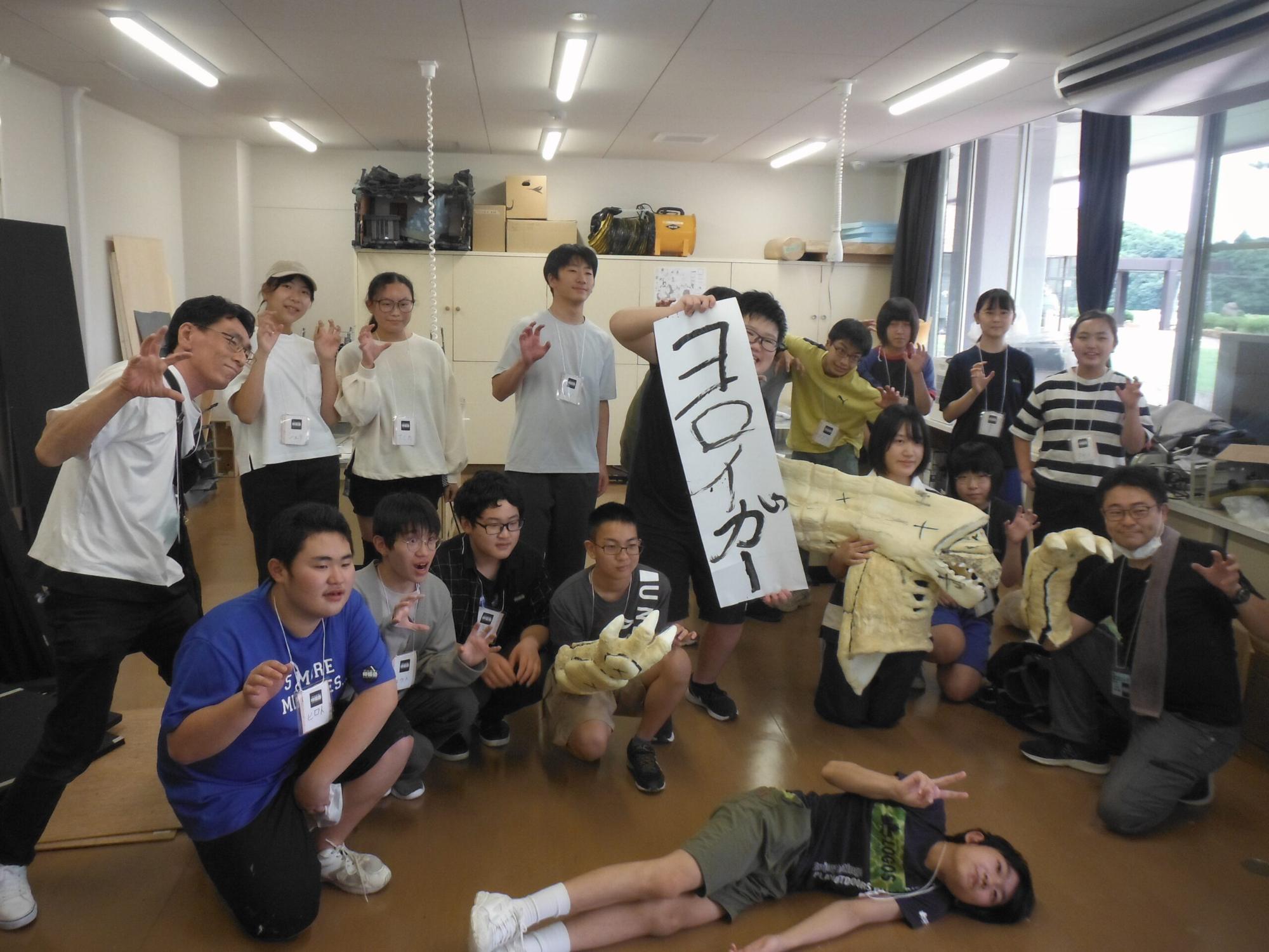 室内で大勢の塾生や講師が集まり、「ヨロイガー」と書かれた紙を手にした塾生や怪獣の工作物を手にポーズを決めている集合写真