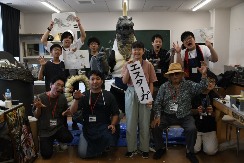 完成した大きな怪獣の着ぐるみを囲み、制作に携わった講師と塾生たちが「エスターガ」と書かれた紙やデザイン画を掲げながらポーズを決めている集合写真