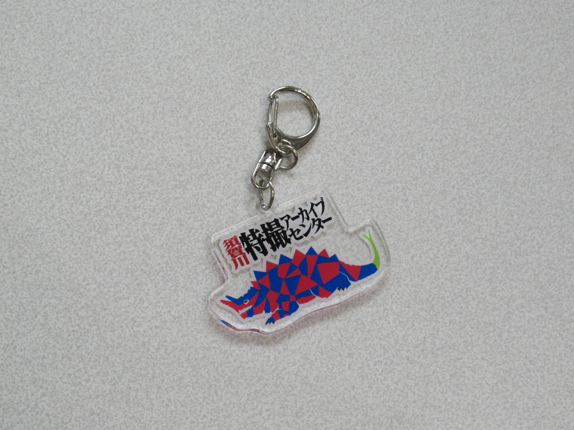 明るいグレーの背景に置かれた、赤と青の幾何学模様で描かれた怪獣のイラストと「須賀川特撮アーカイブセンター」というロゴがプリントされた、透明なアクリル素材のナスカン付きキーホルダーの写真