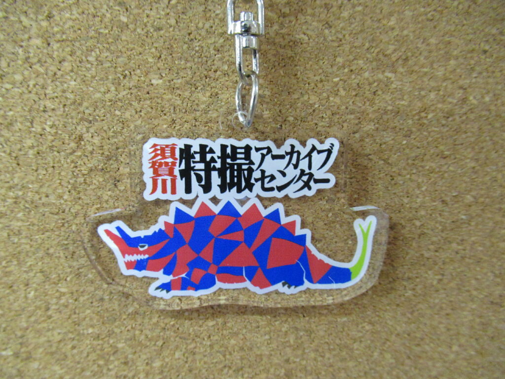 コルクボードを背景に、赤と青の多角形で構成された怪獣のイラストと「須賀川特撮アーカイブセンター」のロゴが描かれた、透明なアクリルキーホルダーを正面から大きく捉えた写真