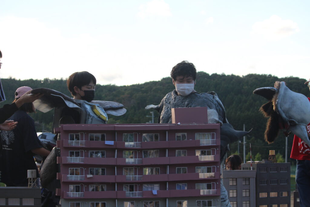 怪獣スーツを着た人物たちがミニチュア建物の背後に立ちポーズや位置を確認しながら特撮演出の準備を進める様子の写真