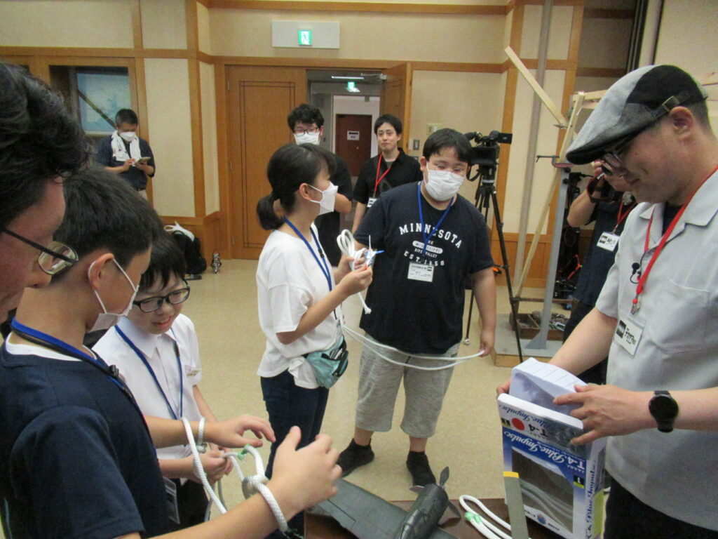 屋内の作業場で、男性が飛行機のプラモデルの箱を手に取り、塾生たちが白い紐を持ち熱心に作業や説明に取り組んでいる様子の写真