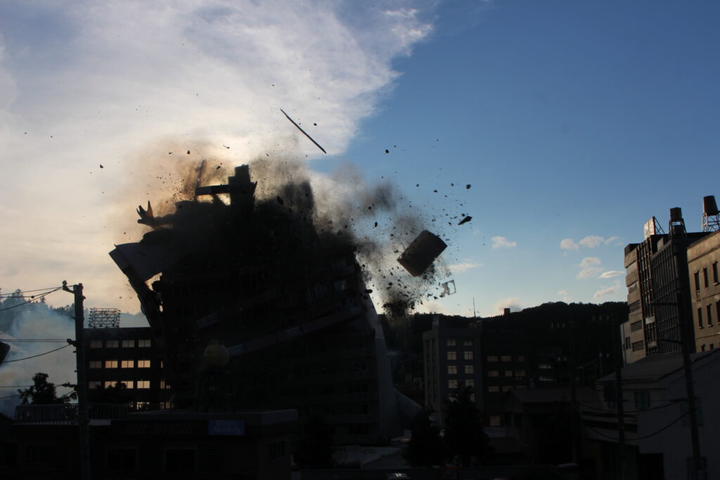 建物模型が激しい爆発の衝撃で大きく崩れ黒煙と無数の破片が四方へ勢いよく飛散し爆炎の広がる迫力ある瞬間を捉えた写真
