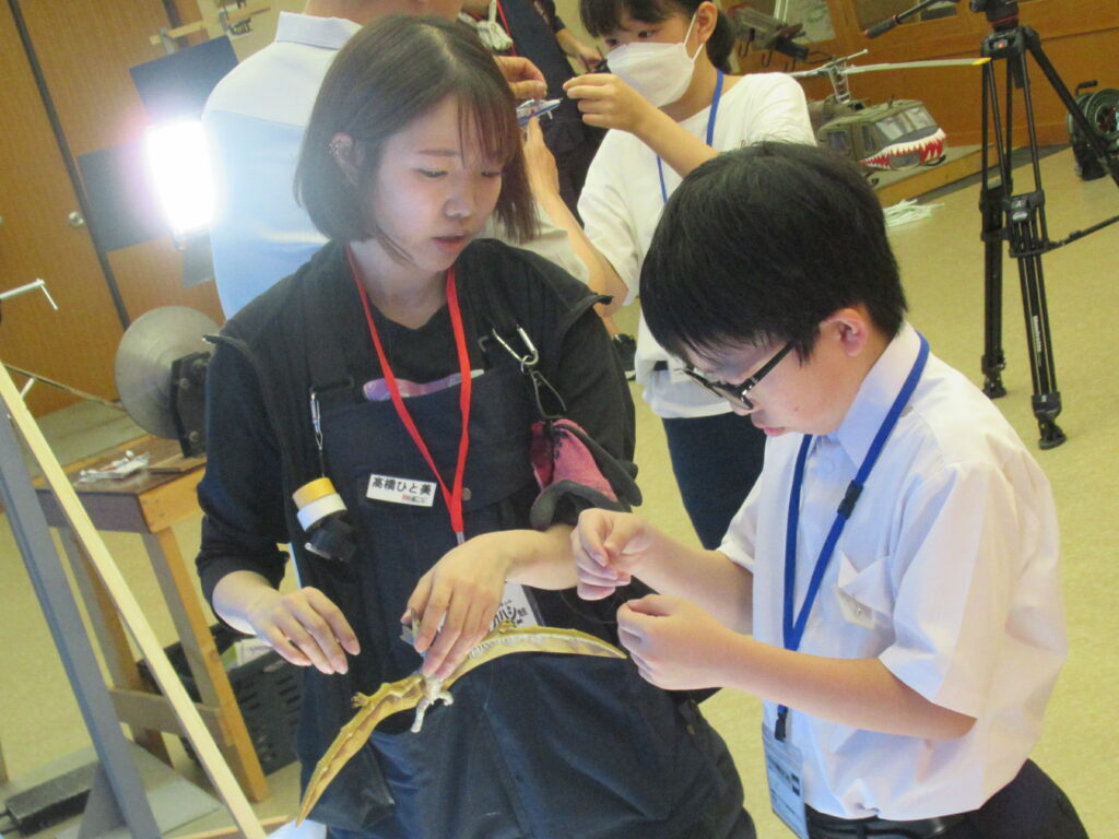 エプロン姿の女性が翼を広げた金色の翼竜のような模型を手に持ち、少年が説明を熱心に聞いている様子の写真