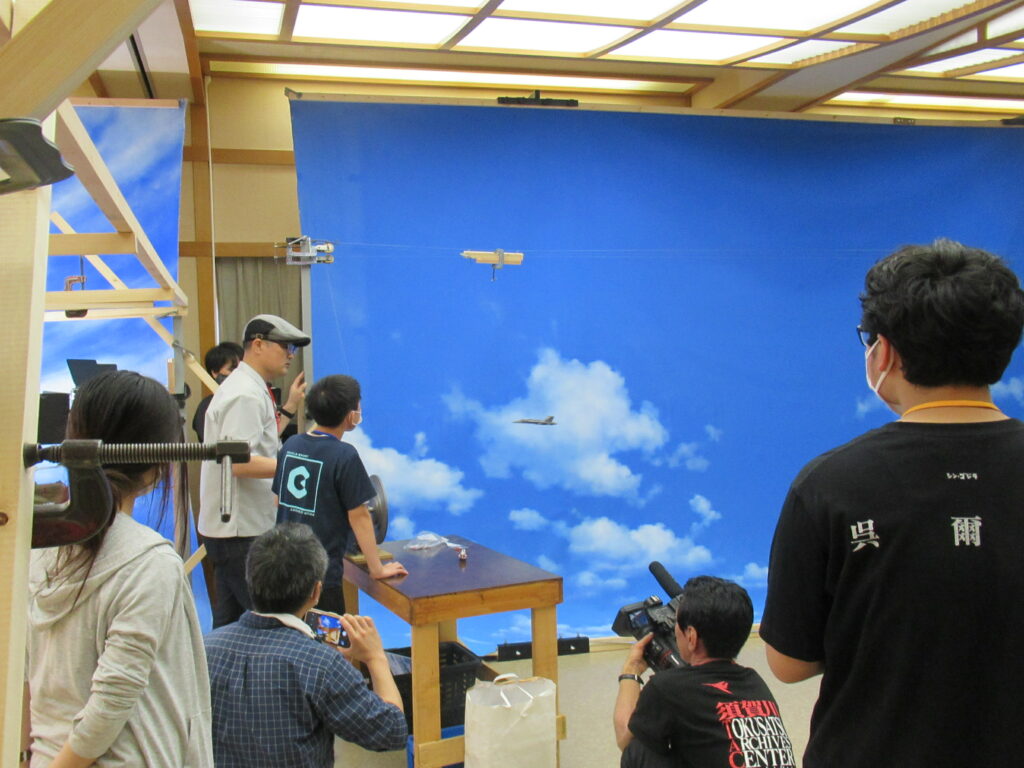 青空の背景スクリーンの前で、ワイヤーで吊るされた戦闘機の模型を飛ばし、手前でカメラを構えるスタッフやそれを見守る参加者たちの写真
