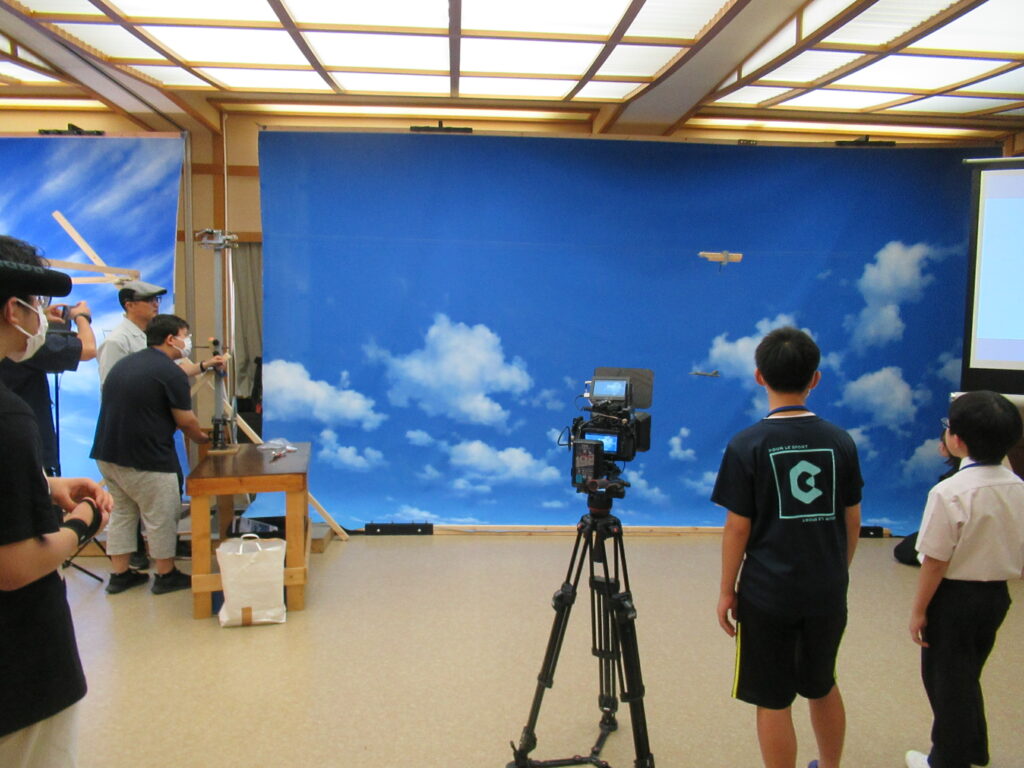 青空と白い雲が描かれた巨大な背景スクリーンの前で、三脚に固定されたカメラを中央に配置し、左側で撮影機材の調整を行うスタッフの姿を見守る塾生の写真