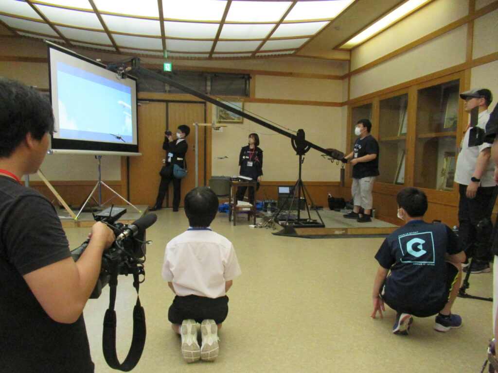 大型のクレーンカメラを操作してスクリーンに映し出された飛行機の映像を撮影しており、手前でひざまずきながらその様子を真剣に見守る塾生の写真
