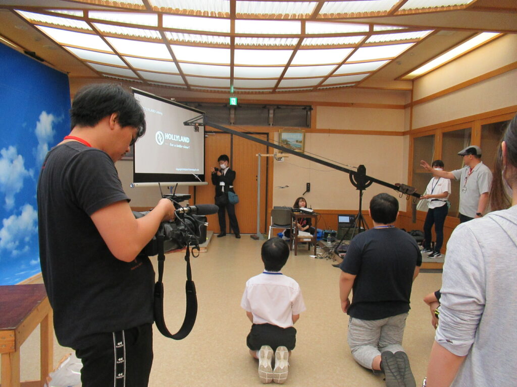 大型のクレーンカメラの操作の説明を真剣に聞いている塾生と手前でひざまずきながらその様子を見守る塾生の写真