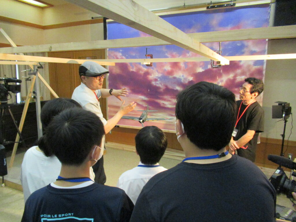 夕焼け空の背景パネルの前で、男性スタッフが身振り手振りを交えて解説し、それを塾生たちが真剣な表情で聞いている写真