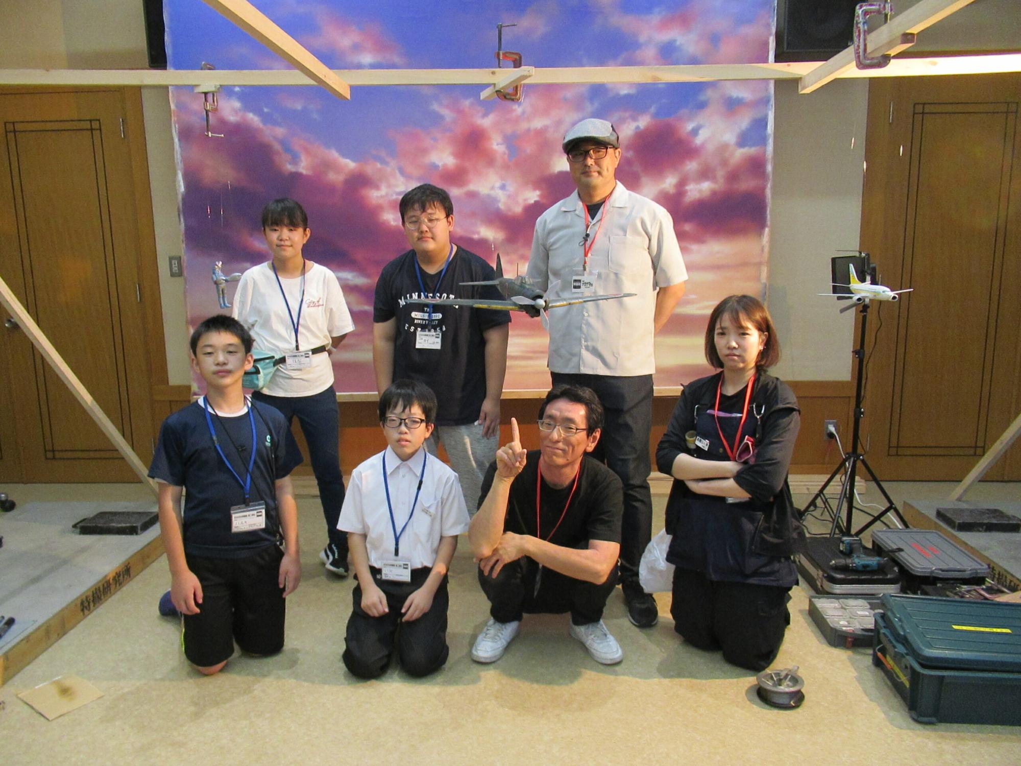 夕焼け空の背景パネルの前に、模型の飛行機を手に持つ男性の横に2人の塾生が並び前列には4人の関係者と塾生がしゃがんで並んで記念撮影をしている写真