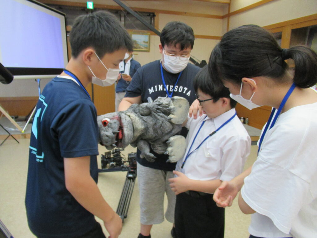 屋内で塾生たちが、ゴジラのような怪獣のパペットに手を入れるのを手伝い確認をしている様子の写真