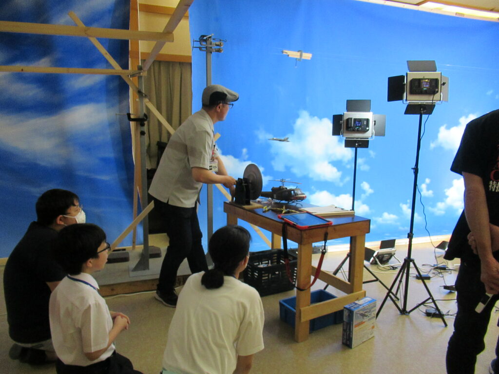 青い空と白い雲が描かれた巨大なスクリーンの前で、照明機材に囲まれながら、スタッフが戦闘機やヘリコプターの模型を使って空中シーンの撮影を進行し、それを手前で塾生たちが興味深く見守っている写真