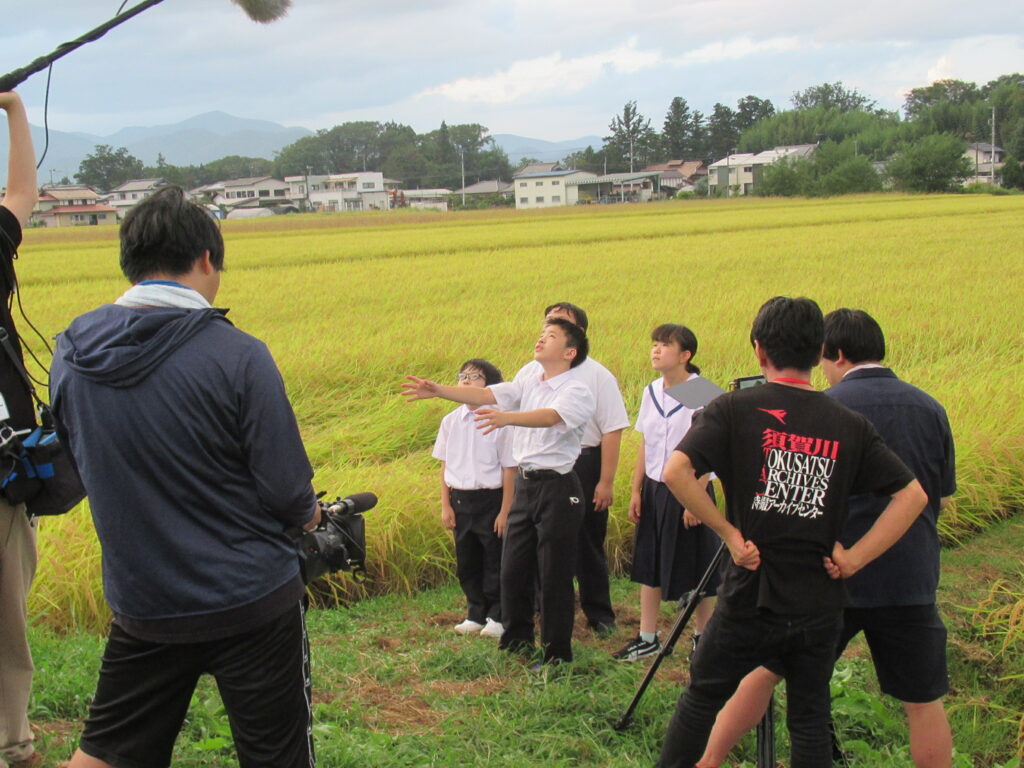 田園風景を背景に、カメラやガンマイクを構えた撮影スタッフの前で、学生服姿の子どもたちが空を見上げてている写真