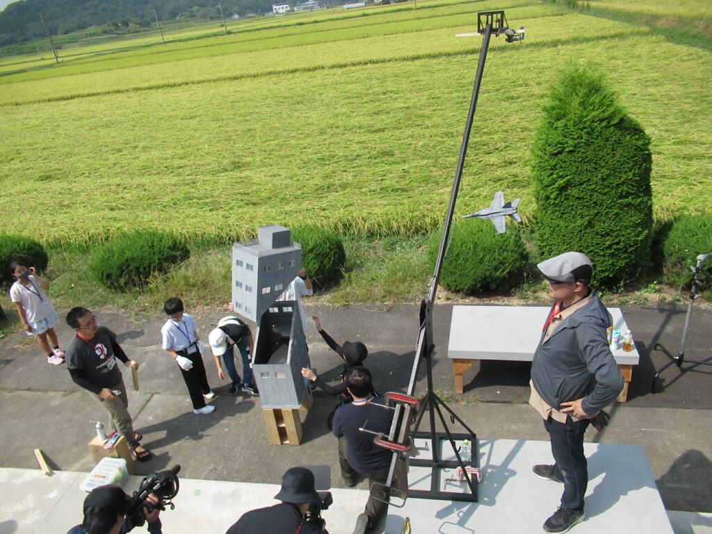田園風景を背景に、屋外で複数のスタッフがミニチュアのビルや長い支柱に吊るされた戦闘機の模型を設置し準備をしている様子の写真