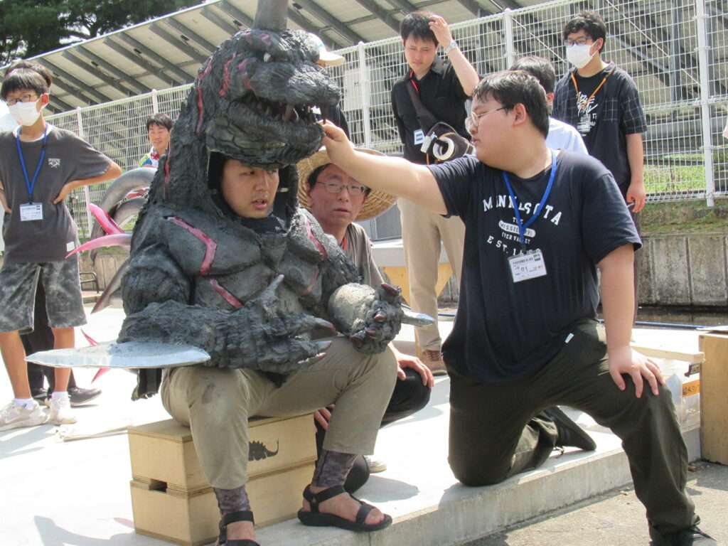 屋外にて、怪獣の着ぐるみを身にまとった男性に講師の話を聞きながら塾生が調整をし準備をしている様子の写真