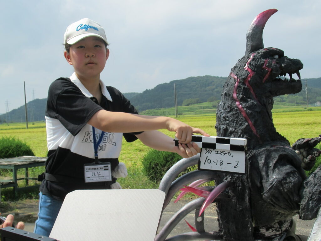 田園風景が広がる屋外で、白い帽子を被った塾生が怪獣の模型の前に立ち、カチンコを掲げて映画撮影の合図を出している様子の写真