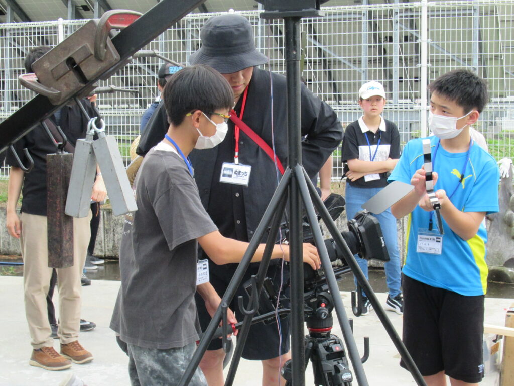 屋外で、三脚に据えられた大きなカメラを囲み、スタッフの指導を受けながら塾生たちが真剣な表情で機材の操作やカチンコの準備を進めている様子の写真