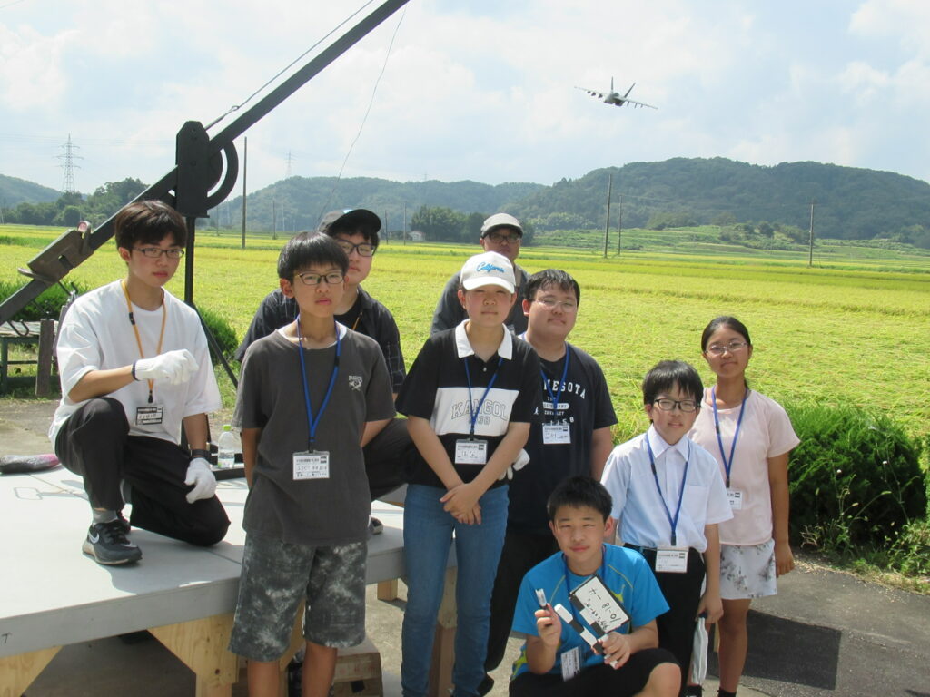 田園風景と山々を背景に、撮影に使用した機材や模型の戦闘機が空に浮かぶセットの前での集合写真