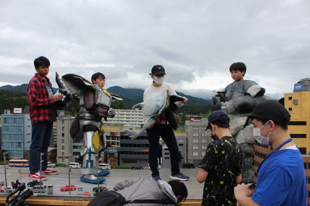 ミニチュア都市模型の上で怪獣スーツを着た人物たちが建物周辺に立ち動きや位置を確認しながら演出準備を進める様子の写真