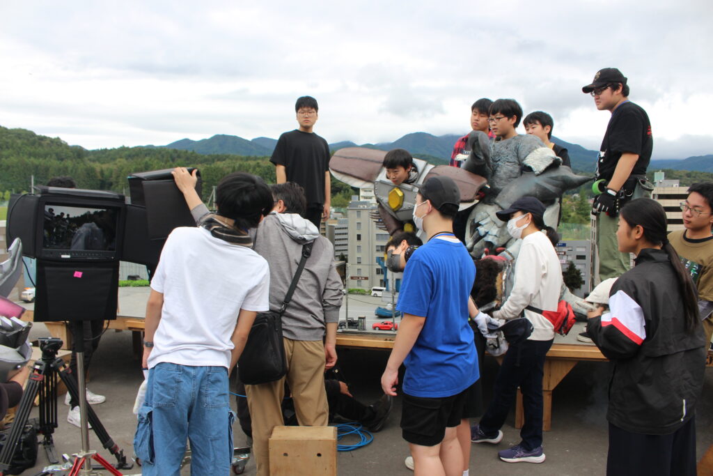 多数の塾生やスタッフがモニター確認や怪獣スーツ調整を行いミニチュア都市模型上で演出準備を進める様子の写真