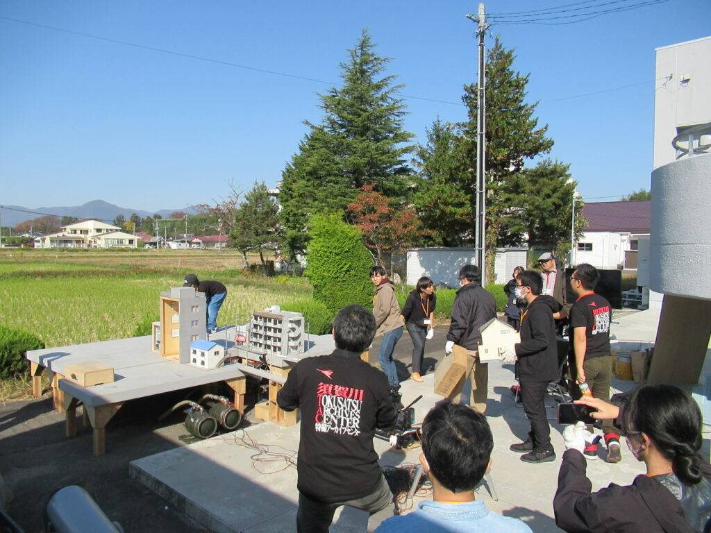 晴天の屋外にて、須賀川特撮アーカイブセンターのロゴ入りTシャツを着たスタッフたちが、台座の上に設置されたビルや住宅などの精巧なミニチュアセットを囲んで撮影の準備や調整を行っている様子を捉えた写真