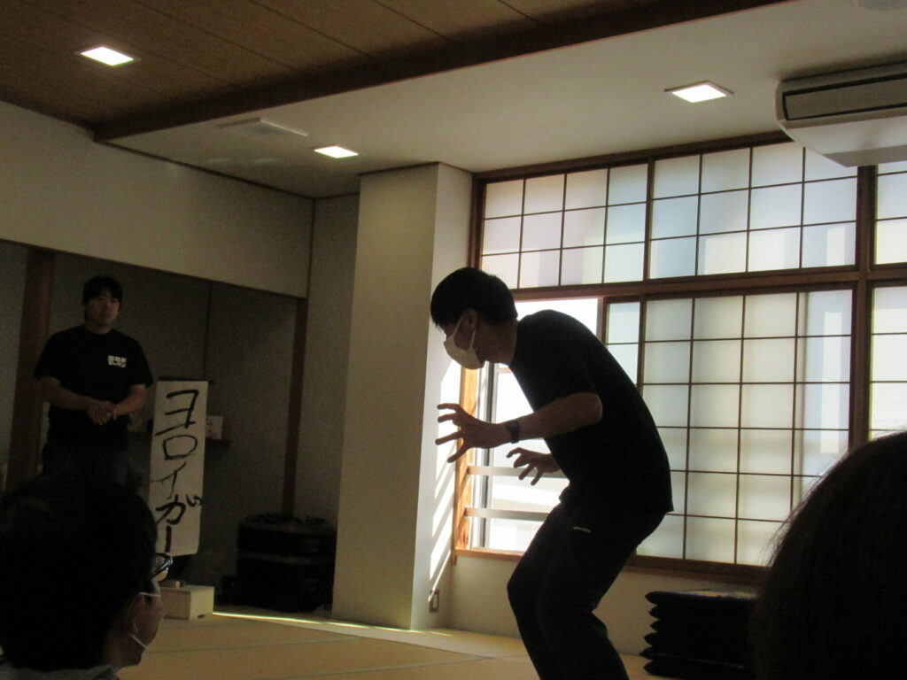 明るい和室の畳の上で、黒いTシャツにマスク姿の男性が、背を丸め両手を鋭い爪のように広げて怪獣のようなポーズを熱演しており、その様子を別のスタッフや観客が真剣な表情で見守っている、演技指導や特撮ワークショップのひとコマを写した写真