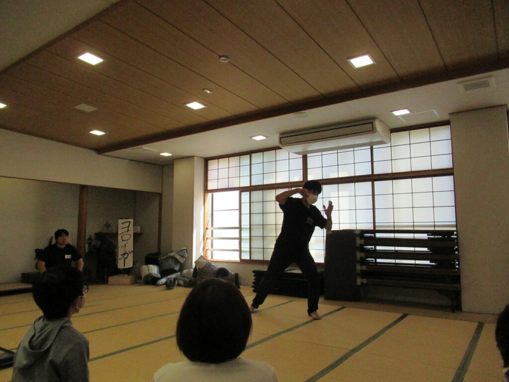 畳が敷かれた和室の広間で、黒いTシャツを着用した男性が窓を背にしてヒーローのような決めポーズを披露しており、それを手前に座った観客たちが静かに見守っている、ワークショップや演劇のワンシーンのような写真