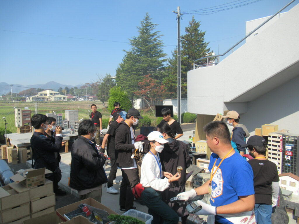 屋外の晴天の下、大勢のスタッフが建物の模型や撮影機材を囲んで特撮の準備作業を行っており、手前には青いTシャツを着た男性が機材を手に持っている写真