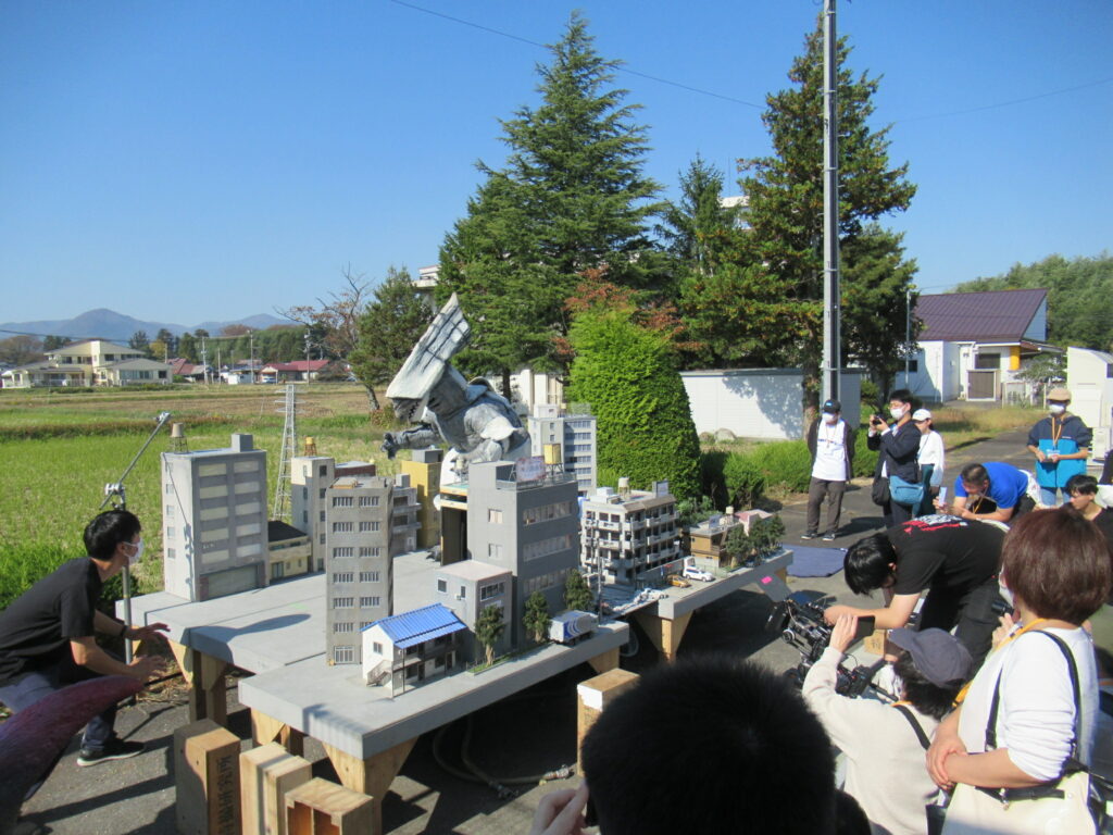 抜けるような青空の下、田園風景を背景に設置された精巧なミニチュアの街並みの中で、巨大なサメのような頭部を持つ銀色の怪獣がビルをなぎ倒すシーンを、大勢のスタッフがカメラや機材を駆使して撮影している臨場感あふれる写真