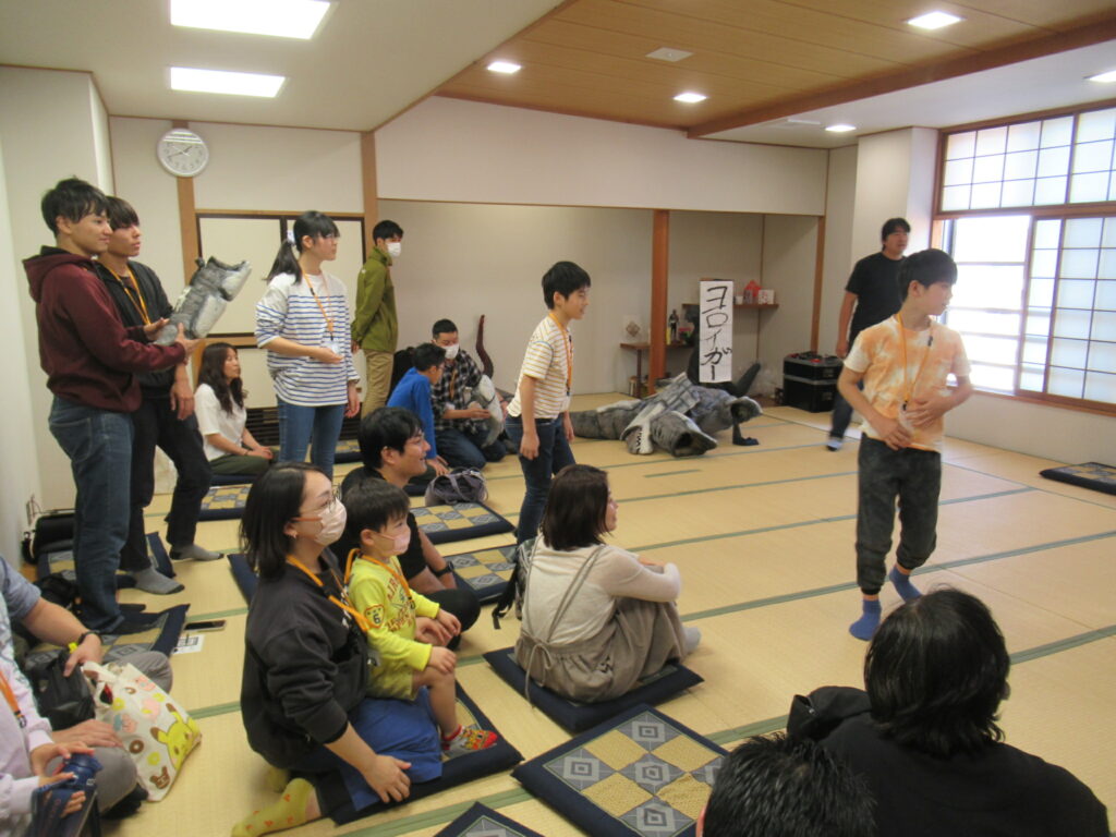 明るい和室の畳の上で、子供たちが演技をする様子を大人や他の子供たちが座布団に座って見守っており、背後には怪獣の着ぐるみや小道具が置かれ、地域や学校のイベントで特撮劇の練習や披露を行っている写真