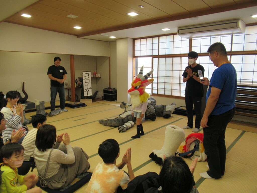 障子の窓がある和室の畳の上で、狐のような頭部を被った子供が立ち、それを取り囲む大人や子供たちが笑顔で拍手を送っており、周囲には怪獣の着ぐるみやパーツが置かれた、特撮体験イベントの締めくくりのような温かな光景を捉えた写真