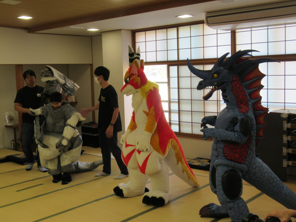 障子の窓がある和室の畳の上で、青い恐竜のような怪獣や赤と白の狐のようなキャラクター、そして岩のような質感の怪獣の着ぐるみを着た人たちが並び、周囲でスタッフがサポートしながら撮影や演技の準備を進めている写真