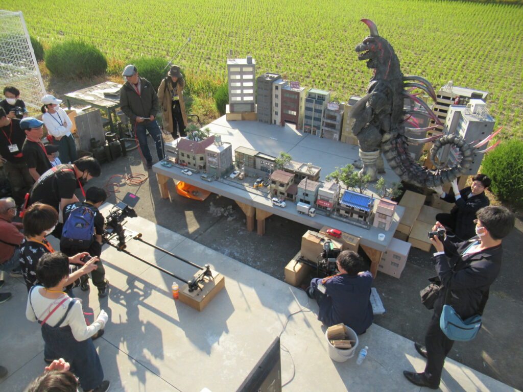 晴れた屋外で、田園風景を背に設置されたミニチュアの街並みのセットを囲み、大きな怪獣の模型を動かしたりカメラを構えたりして、特撮映画のような撮影を熱心に行っている大勢のスタッフたちの様子を俯瞰で捉えた写真