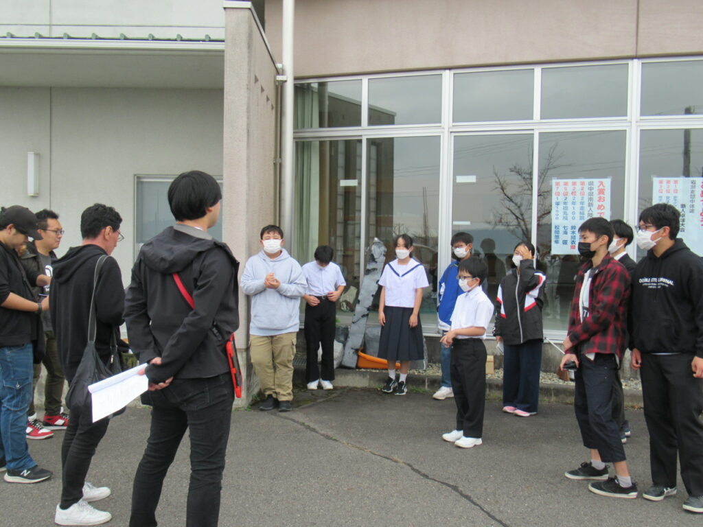 建物の外で、台本のような紙を手にした制作スタッフと、制服や私服姿の生徒たちが集まり、撮影の打ち合わせの説明を聞いている写真