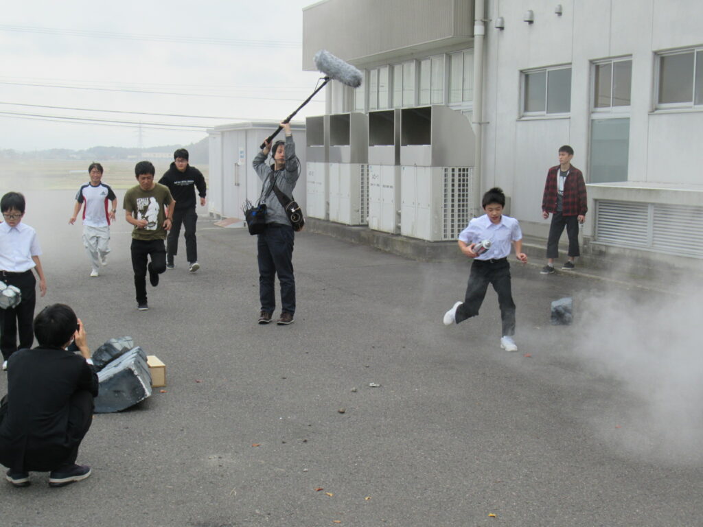 屋外の舗装された場所で、白煙が上がる中を制服姿の生徒や私服姿の生徒が必死に駆け抜け、その様子を音声スタッフが長いマイクを掲げながら撮影している写真