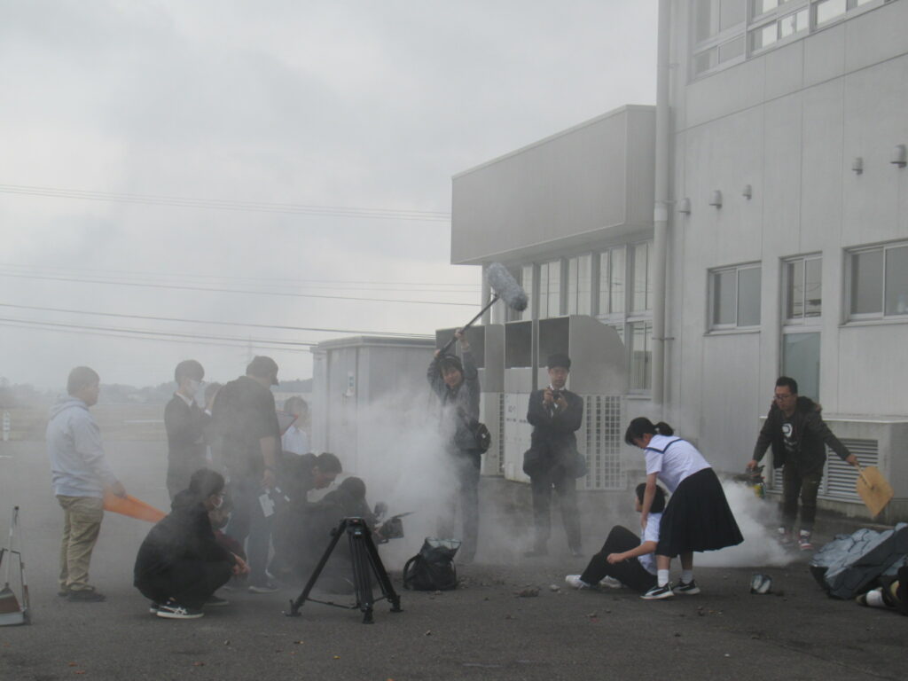 激しい白煙が立ち込める建物の外で、倒れ込む生徒を助けようとする生徒の姿を、多くのスタッフが撮影している様子の写真