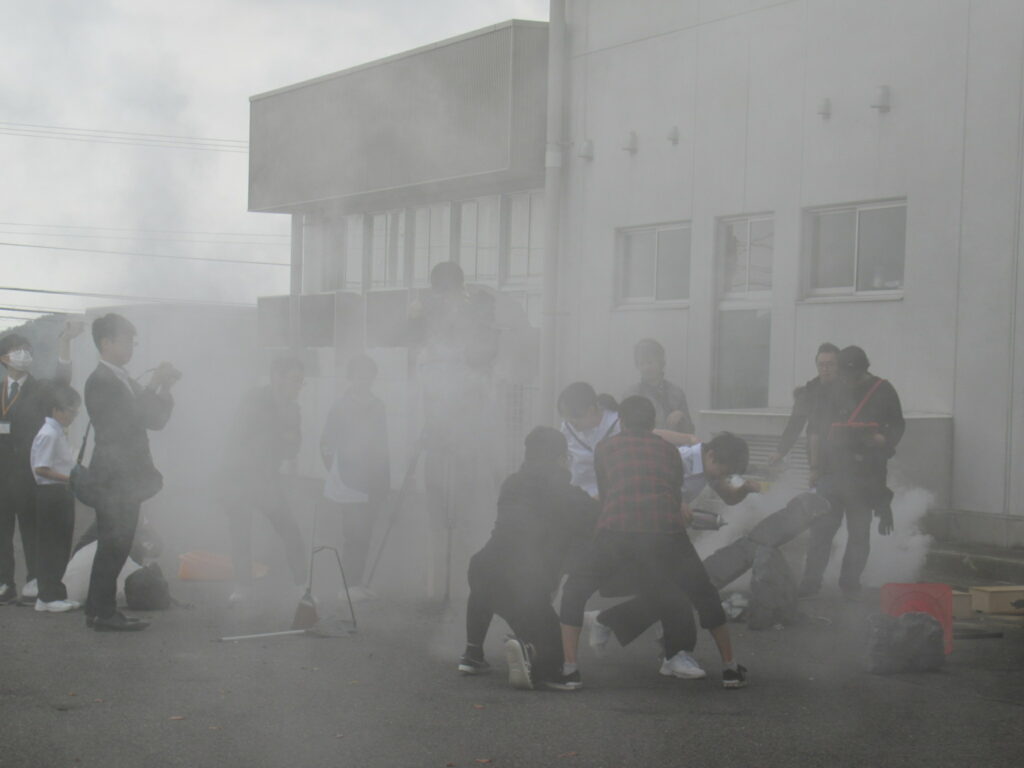 視界が遮られるほどの濃い白煙に包まれた屋外の撮影現場で、スタッフや生徒たちが一点に集まり、機材を操作したり演技をサポートしたりしながら一丸となって映画制作に取り組んでいる、熱気あふれる制作現場の写真