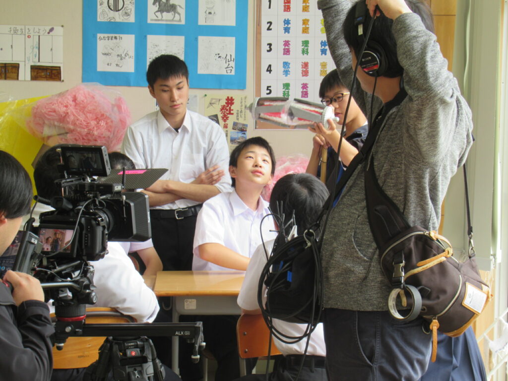 教室内でカメラや録音機材を構えるスタッフに囲まれ、制服姿の生徒たちが真剣な表情で演技に臨んでいる様子の写真
