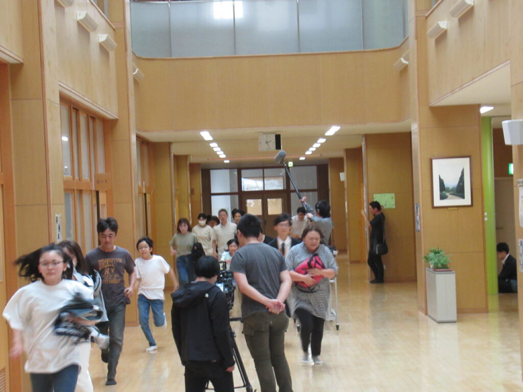 吹き抜けのある広々とした校舎の廊下で、多くの方々が逃げ惑うような演技をしながら一斉に走り出し、その様子をカメラやマイクで撮影している写真