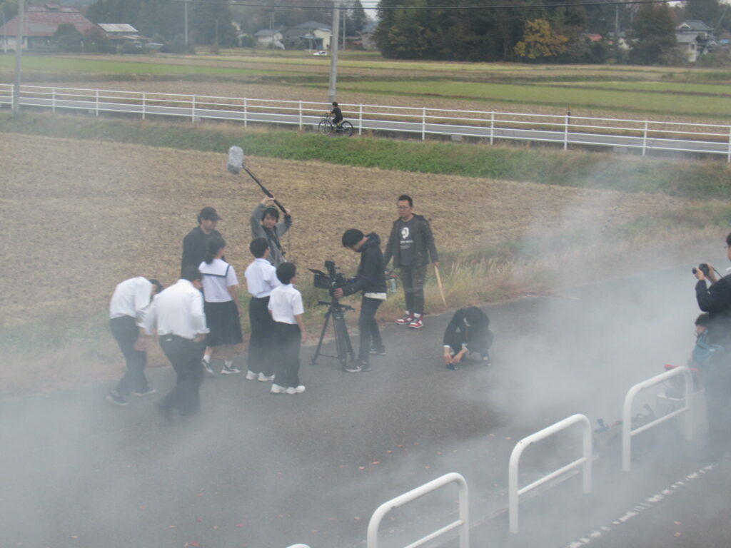 田園風景が広がる屋外の道で、白煙を漂わせながら、制服姿の生徒たちやカメラ・音声機材を構えたスタッフが一点に集まって撮影を進めている写真