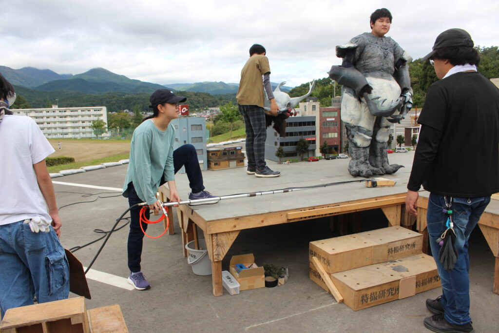 ミニチュア都市模型の上で怪獣スーツの男性とスタッフが位置や動作を確認し撮影準備を進める様子の写真