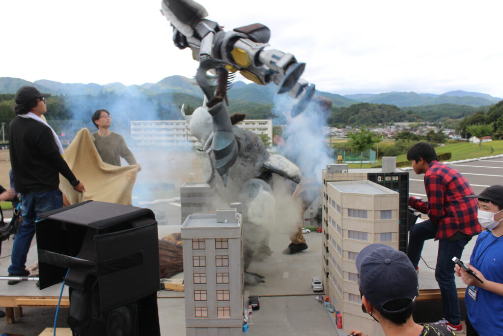 怪獣スーツの人物がミニチュア都市の中央で腕を振り上げ煙の中ポーズを取り周囲のスタッフが動作確認を行う様子の写真