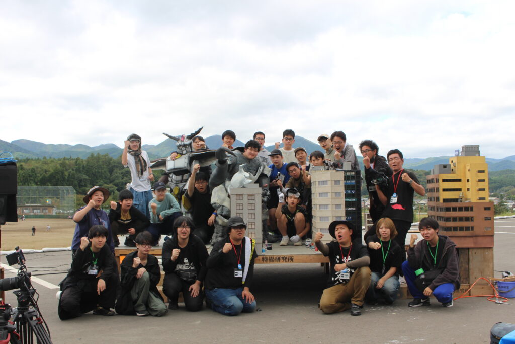 多数の参加者がミニチュア都市模型の周囲で怪獣スーツとともに拳を突き上げ記念撮影に臨む様子の写真