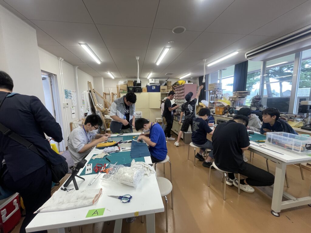 作業室で複数の塾生がカッターマット上で裁断や組み立て作業に集中し周囲でも道具や素材を扱いながら制作工程を進めている様子の写真
