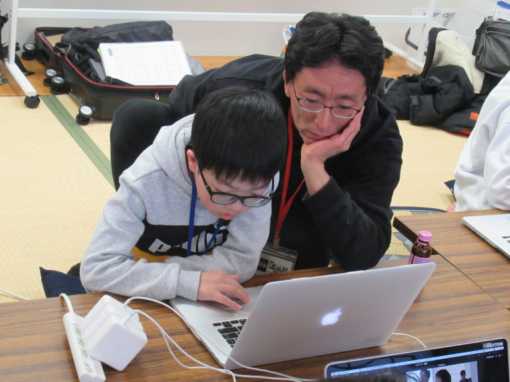 和室の長机でノートパソコンを操作する少年の傍らに男性の講師が寄り添い、二人で真剣な表情を浮かべながら画面に映る編集中の映像を確認している様子の写真