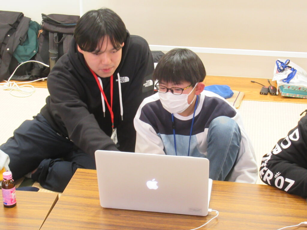 和室に座る少年の横に男性講師が並び、ノートパソコンの画面を指し示しながら、映像編集の説明をしている様子の写真