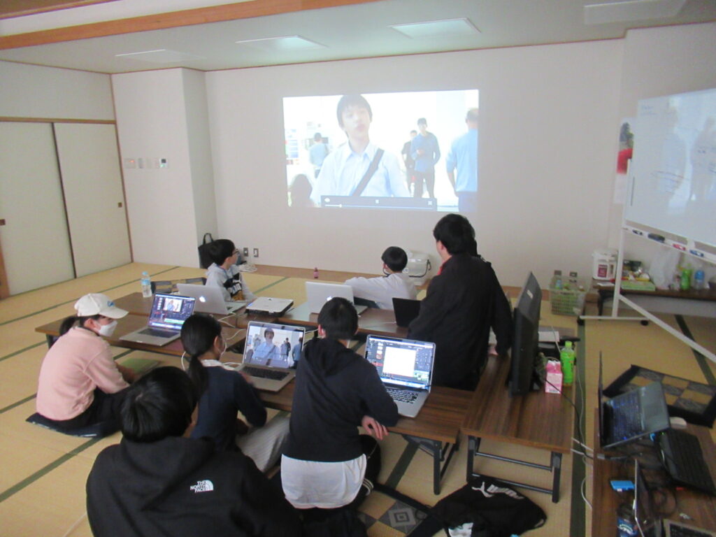 和室の白い壁面にプロジェクターで映像が投影され、座卓を囲んだ参加者たちが自身のノートパソコンを開きながら、映し出された動画を見つめている写真