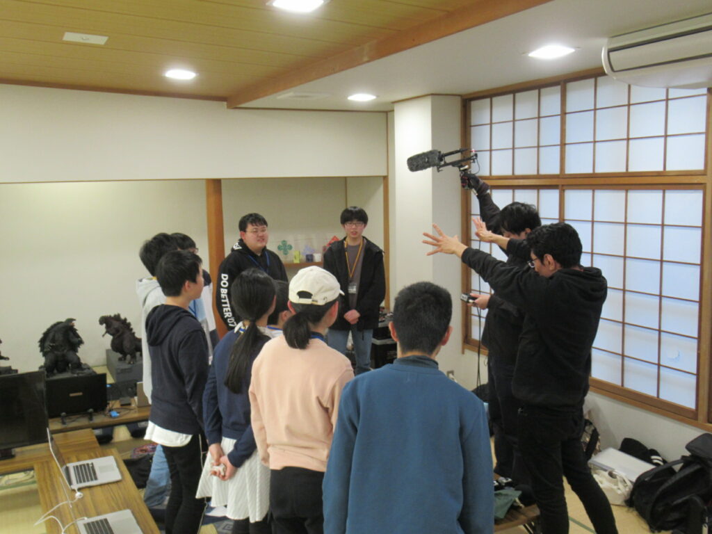 和室で講師が、ガンマイクを高く掲げ、両手を広げて説明をし、それを取り囲む若者たちが真剣な表情で見守っている写真