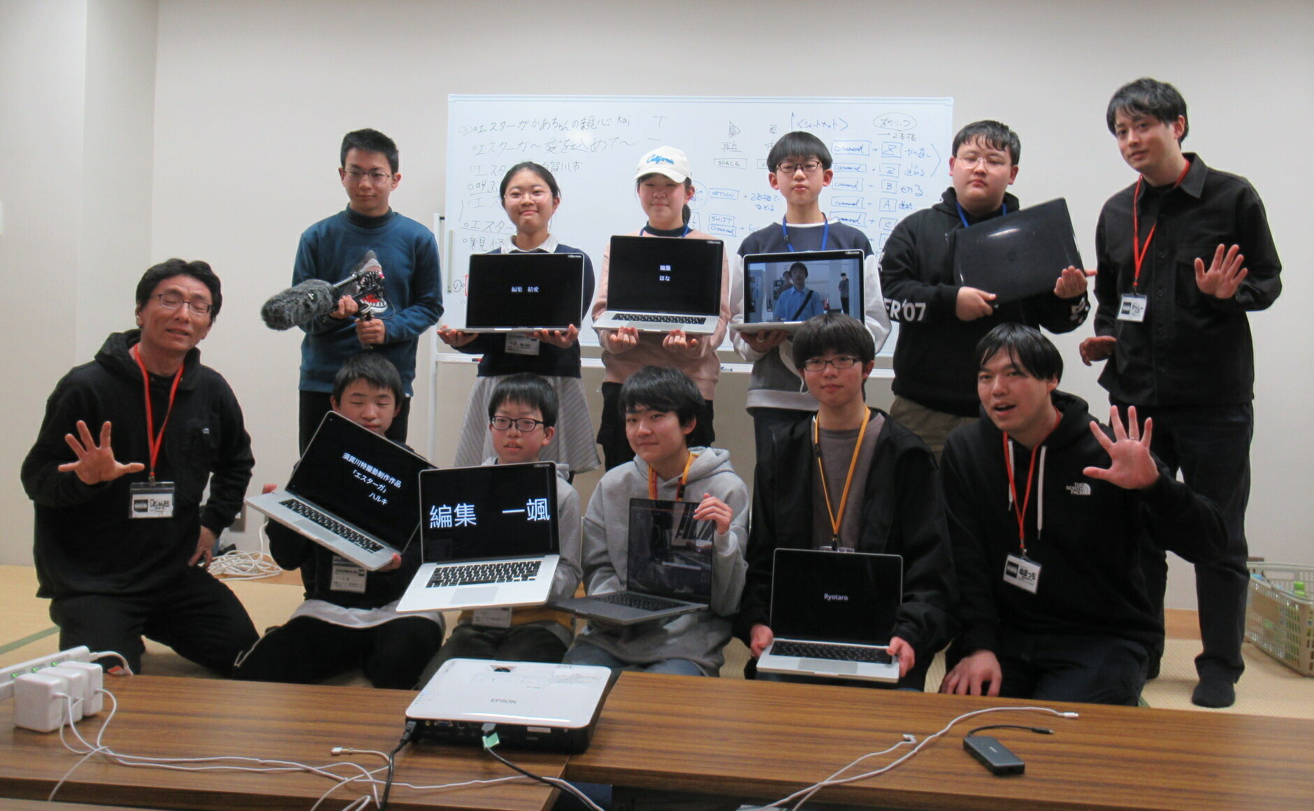 ノートパソコンを手にした複数の参加者たちと講師の男性たちが二列に並び、画面に表示された文字や映像を見せながら記念撮影している写真